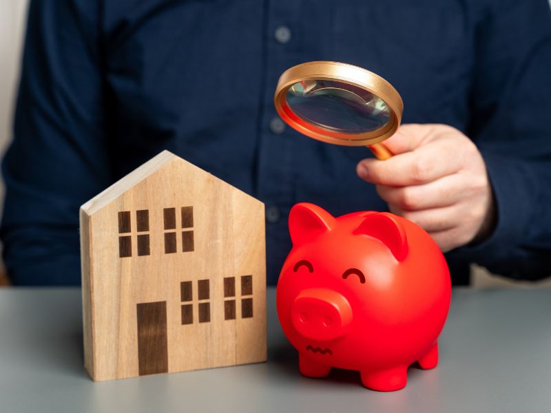 osoba drži lupu u ruci, pored kasica prasica i kuća